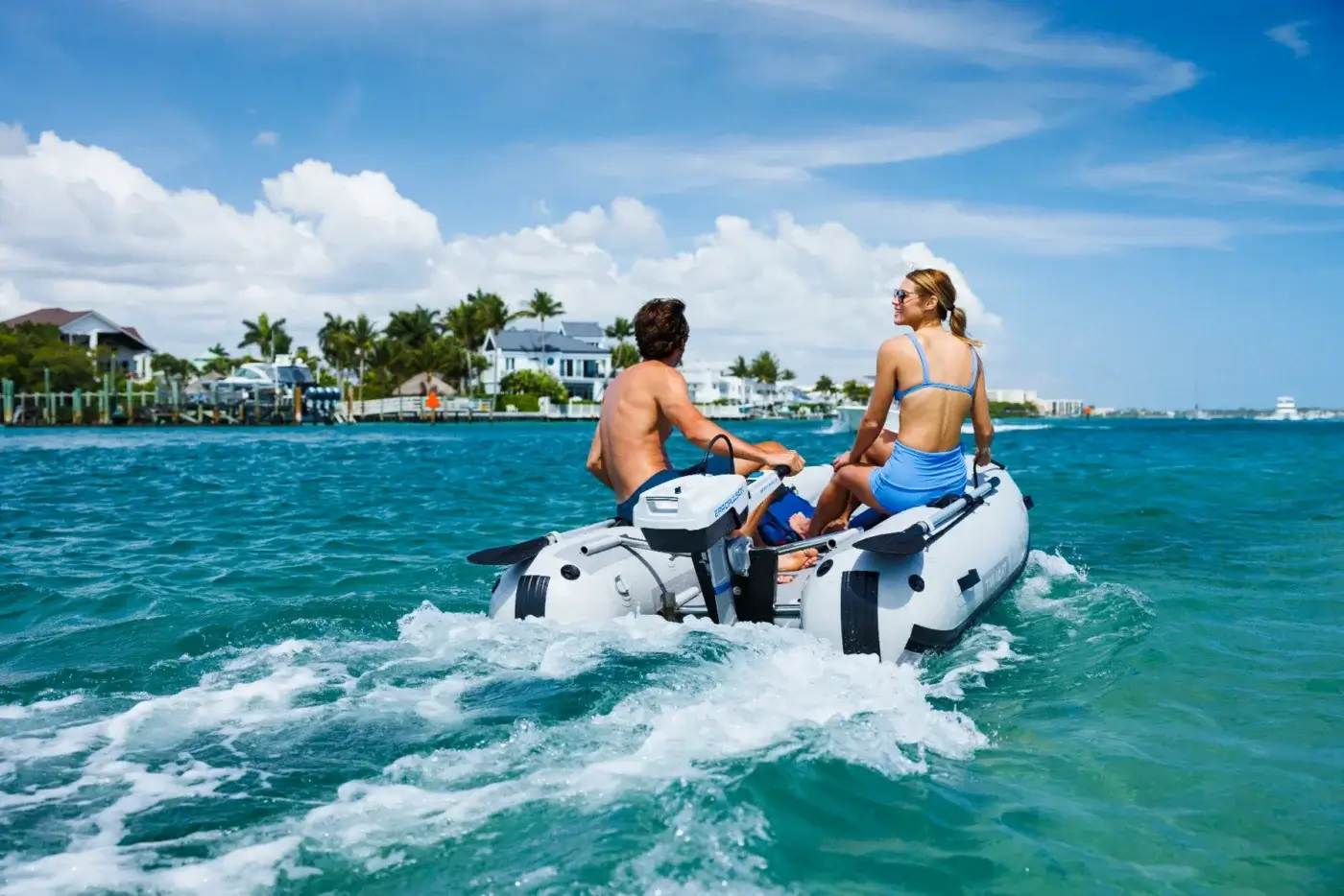 Two people riding an inflatable motorboat with an electric outboard motor on a sunny day with waterfront homes in the background.