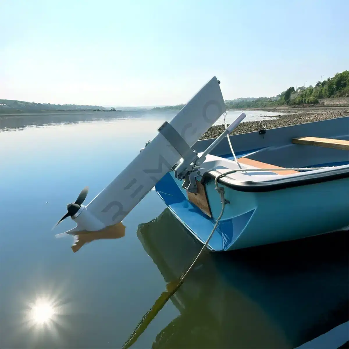 A small blue rowboat with a white electric motor attached at the stern is moored in calm water near a rocky shoreline, with trees and hills visible in the background under a clear sky. Oceans EV