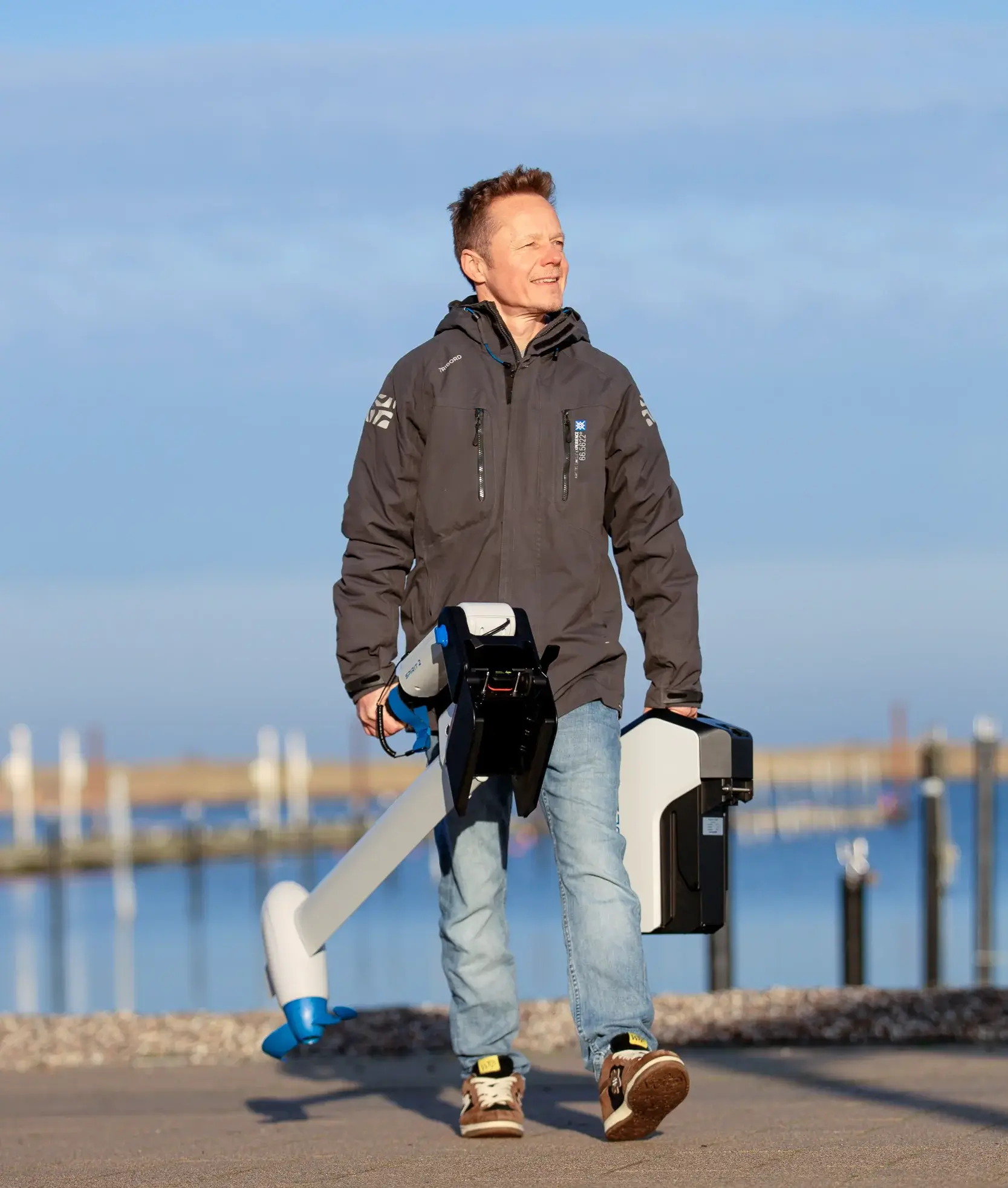 A man in a gray jacket and jeans walks outdoors near water, smiling and carrying a large piece of equipment with blue and white parts in one hand and a black case in the other. The background shows docks and calm water. Oceans EV